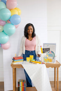 Full table scene with woman standing behind scalloped-edge wood table. Crayon boxes in yellow and mint at center. Books stacked to the left, a small school milk carton visible among props. Grid table runner, white shiplap backdrop.