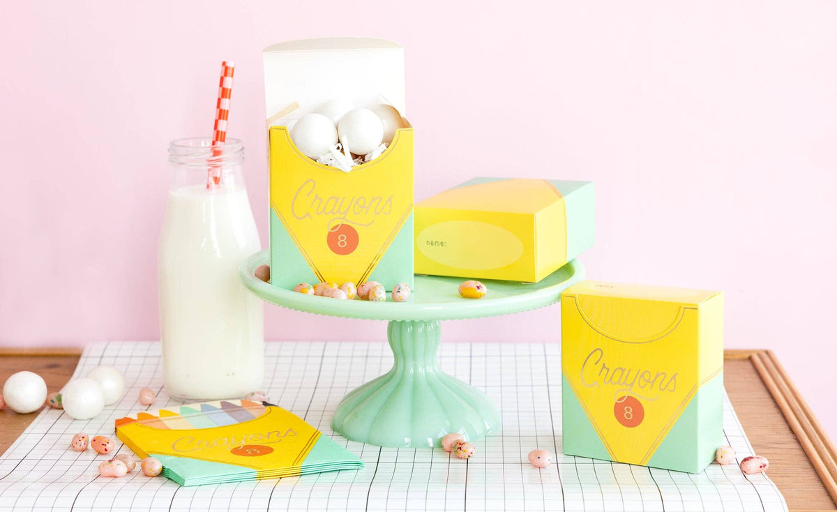 Closer version of Image 17 minus the tipped box. Open crayon box on cake stand holds white cake pops; closed yellow name-label box sits beside it; standing crayon box at right shows "Crayons 8" script. Milk bottle with red straw to left. Candy scattered on runner.