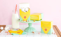 Closer version of Image 17 minus the tipped box. Open crayon box on cake stand holds white cake pops; closed yellow name-label box sits beside it; standing crayon box at right shows "Crayons 8" script. Milk bottle with red straw to left. Candy scattered on runner.