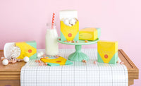 Wide shot of a styled dessert table on a grid-print white runner against a pink wall. Assembled crayon treat boxes used as favor boxes — one tipped on its side with white crinkle paper and marshmallow balls spilling out, one upright on a mint green cake stand holding white cake pops, one standing beside a glass milk bottle. A second style of yellow box (name label on front) sits on the cake stand. Stack of napkins in foreground.