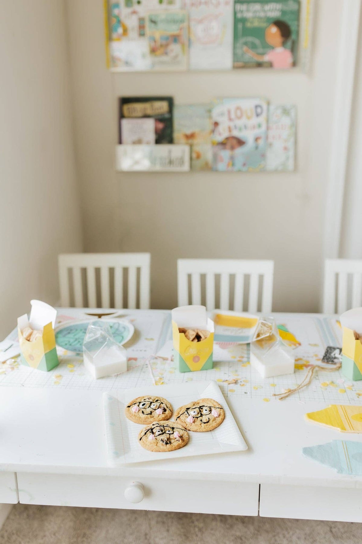 White kids' table with four chairs in a reading nook. Assembled crayon treat boxes used as individual place settings — open and holding animal crackers, one at each place. Globe plate, flat crayon-print napkins, and confetti visible. Picture books on wall ledge in background.