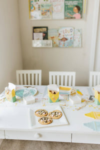White kids' table with four chairs in a reading nook. Assembled crayon treat boxes used as individual place settings — open and holding animal crackers, one at each place. Globe plate, flat crayon-print napkins, and confetti visible. Picture books on wall ledge in background.