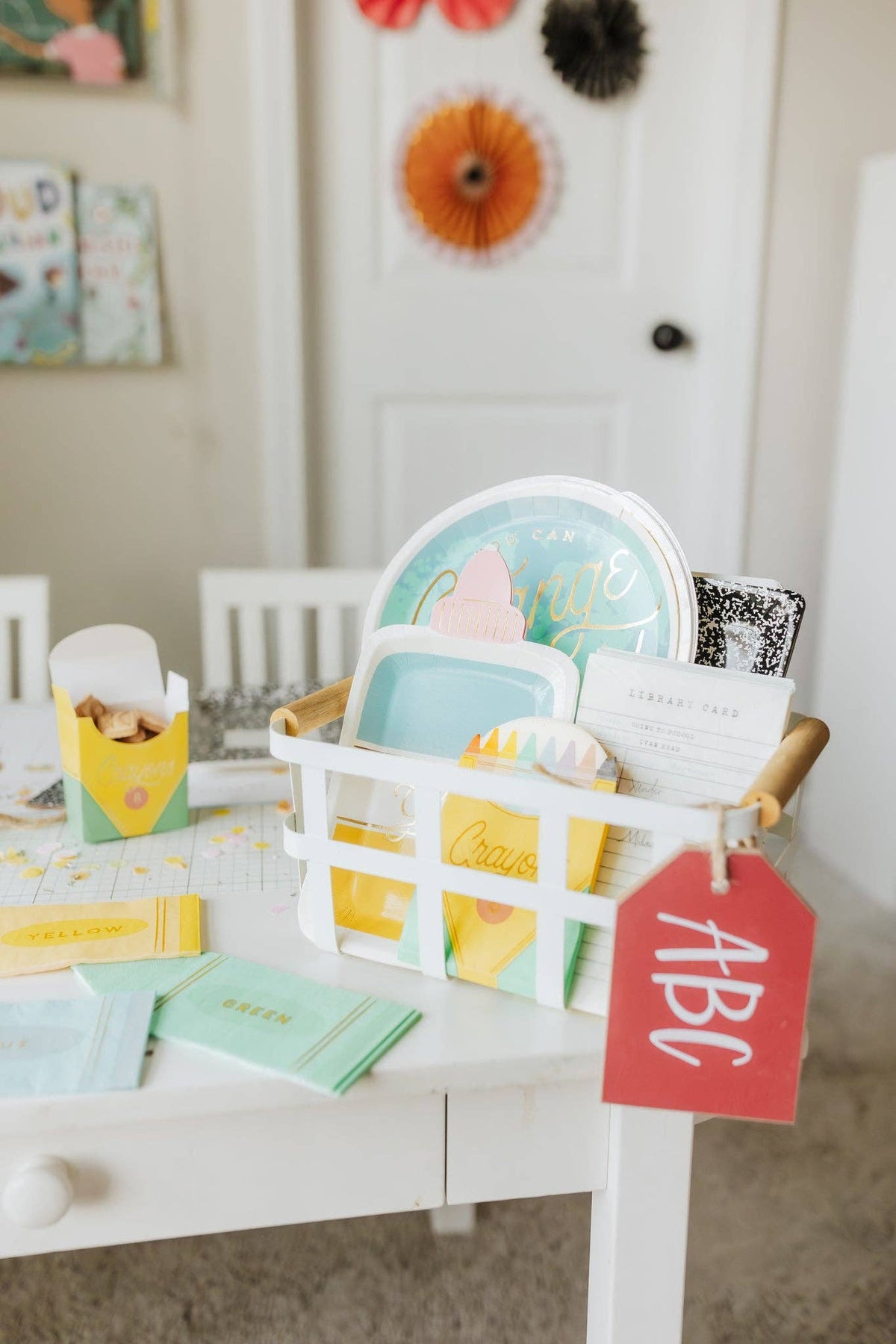 Close-up corner of the kids' party table showing a white wire basket holding the globe plate, composition notebook plate, library card pad, and folded crayon-print napkins labeled "Yellow" and "Green." Crayon treat box visible left. Red ABC tag hangs from basket handle.
