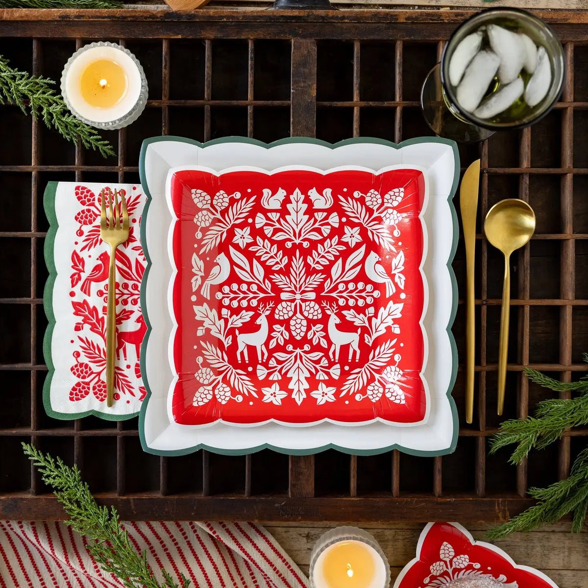 Cream square paper plate with a green scalloped edge under a red woodland folk art plate on a vintage type tray with a woodland napkin and gold flatware