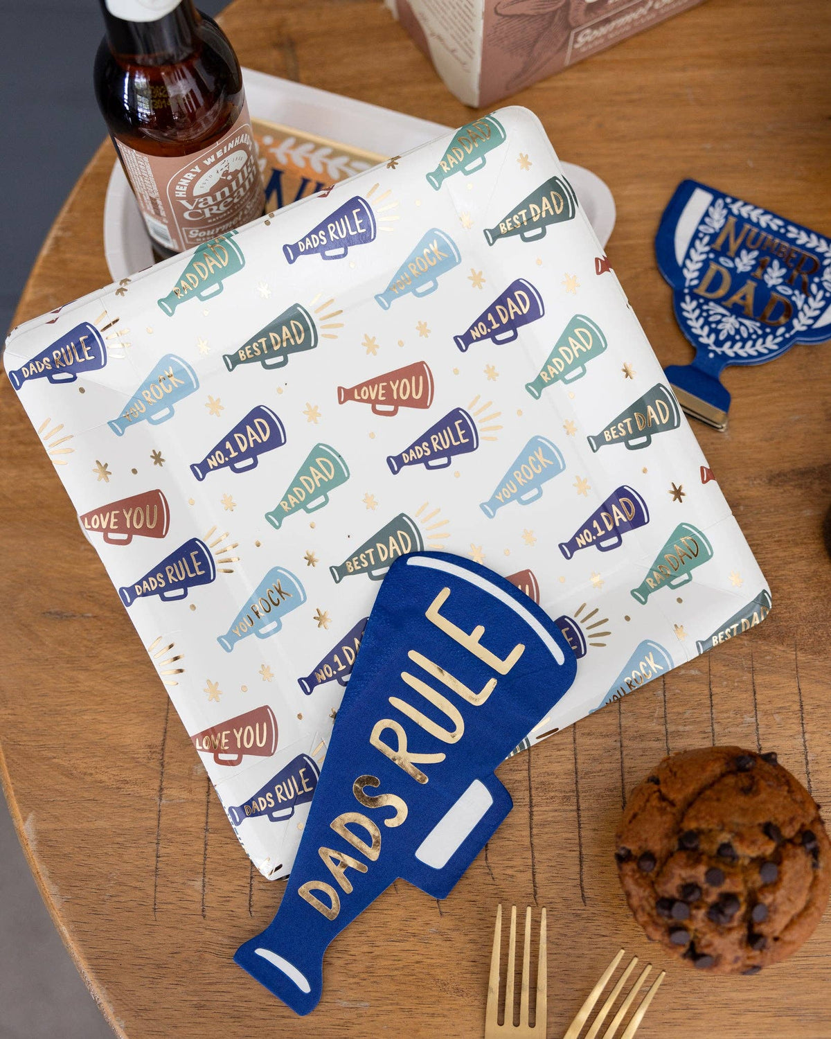 Father's Day tablescape with Dads Rule megaphone paper cocktail napkin on a white square paper plate printed with multi-color megaphones reading "Best Dad," "Rad Dad," "No. 1 Dad," "Love You," and "Dads Rule," styled with Number 1 Dad trophy napkin, vanilla cream soda bottle, gold forks, and a chocolate chip cookie