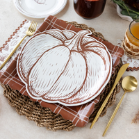 An angled view of a fall table setting with a pumpkin-shaped plate on a brown plaid placemat and woven charger.