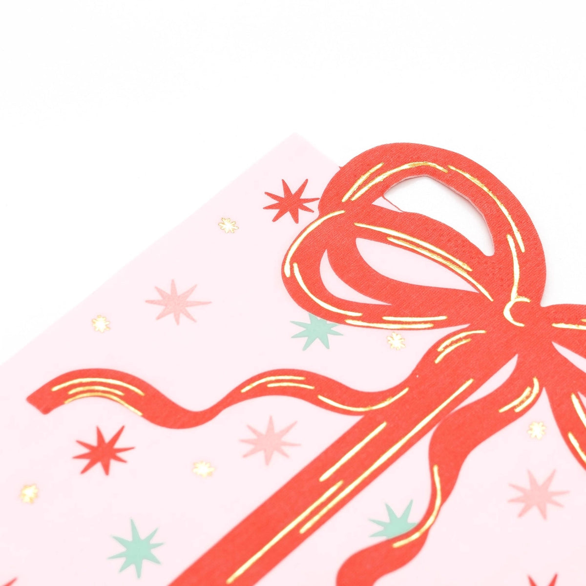 Close-up macro view of the die-cut red ribbon bow and gold foil details on the top half of the pink holiday napkin.