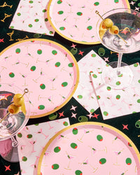 Overhead view of pink martini print paper plates with slices of pink cake on pink background showing martini glass and olive pattern with gold foil rim