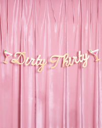 Full Dirty Thirty gold foil script banner with martini glass accents displayed against a pink velvet draped backdrop
