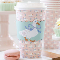 A lifestyle photo of a pink duck to-go coffee cup on a marble table next to a pink macaron and spring cupcakes for a garden party.