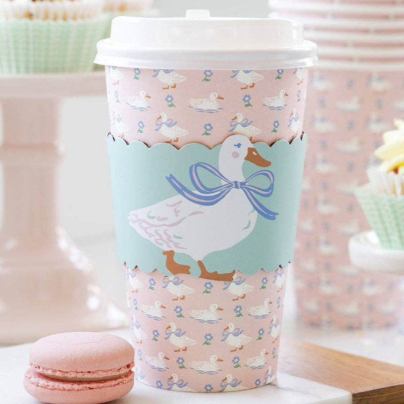 A lifestyle photo of a pink duck to-go coffee cup on a marble table next to a pink macaron and spring cupcakes for a garden party.