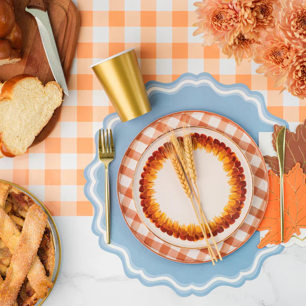 Overhead fall tablescape on an orange gingham paper runner. Dusty blue scalloped placemat under a stack of orange plaid and sunflower-border ceramic plates. Gold paper cup, gold fork, fall leaf-shaped napkins, fresh orange mums, and a bread board with rolls visible around the setting.