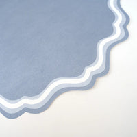Close-up corner shot of a single dusty blue paper placemat showing the scalloped die-cut edge and double-wave concentric border detail — outer ring in medium blue, center stripe in white, inner ring in lighter blue. Matte paper texture visible.