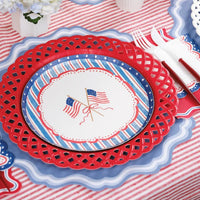 Close-up of a single July 4th place setting on a red striped tablecloth. Dusty blue placemat under a red lattice-cut charger plate, then a crossed-American-flag scalloped dinner plate. White plastic fork and knife in a napkin roll to the right.