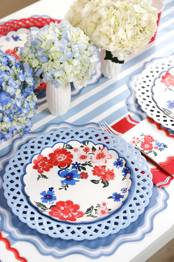 Angled shot of the same patriotic tablescape. Single foreground place setting shows the layered look: blue scalloped placemat → white lattice charger → red floral plate. Red-striped napkin with gold fork. Blue hydrangeas and white hydrangeas in milk jugs down the center of the table.