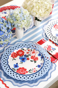 Angled shot of the same patriotic tablescape. Single foreground place setting shows the layered look: blue scalloped placemat → white lattice charger → red floral plate. Red-striped napkin with gold fork. Blue hydrangeas and white hydrangeas in milk jugs down the center of the table.