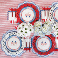 Overhead of a full July 4th table with red striped tablecloth. Each place setting alternates dusty blue and red scalloped placemats under white lattice chargers and crossed-flag scalloped paper plates. White flatware, red folded napkins, white daisies and blue hydrangeas as centerpieces.