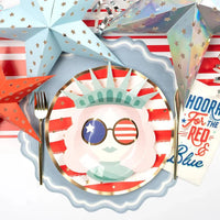 Overhead single place setting on a white surface. Dusty blue scalloped paper placemat underneath a Statue of Liberty illustrated paper plate with red-and-white stripes and gold foil border. Gold fork to left. Iridescent star decorations and "Hooray for the Red, White and Blue" napkin in background.