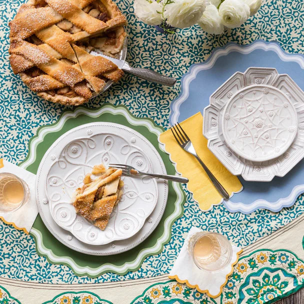 Overhead shot of two place settings on a teal block-print floral tablecloth. Dusty blue placemat (foreground) and green scalloped placemat (background) each under white embossed ceramic plates. Apple pie with a slice cut and a gold fork in place. Yellow linen napkins. Champagne glasses at lower right.