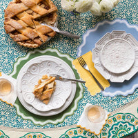 Overhead shot of two place settings on a teal block-print floral tablecloth. Dusty blue placemat (foreground) and green scalloped placemat (background) each under white embossed ceramic plates. Apple pie with a slice cut and a gold fork in place. Yellow linen napkins. Champagne glasses at lower right.