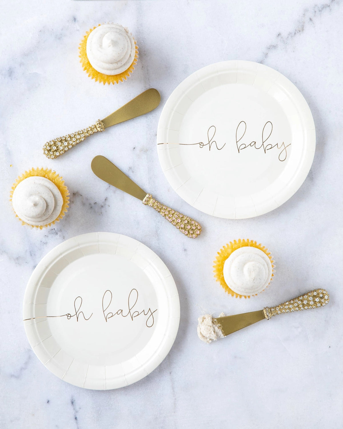 Flat lay of “oh baby” paper plates on marble surface with cupcakes and gold serving knives