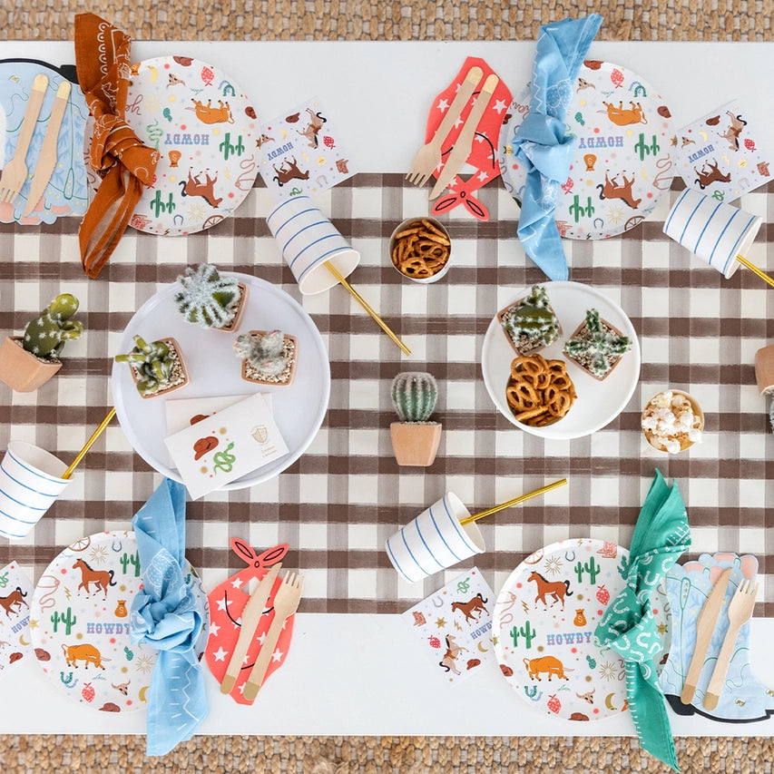 Overhead view of a checkered table runner set with red bandana‑shaped napkins, cactus decor, and mini western plates.