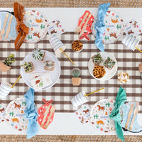 Overhead view of a checkered table runner set with red bandana‑shaped napkins, cactus decor, and mini western plates.