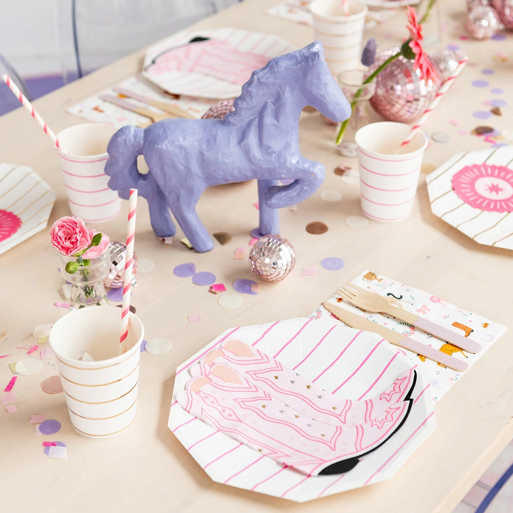 Overhead shot of a pale-wood table set for a cowgirl gathering: pink boot napkins on striped plates, horse-print napkins, pink disco balls, and wood-fork place settings.