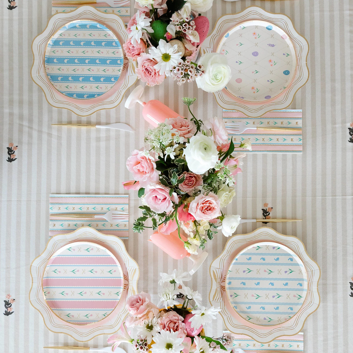 Overhead Easter brunch table with four pastel plate designs, pink vases, gold cutlery and pink floral centerpiece on striped linen