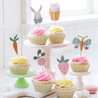 A tiered cake stand displaying cupcakes with pink and yellow frosting, decorated with bunny, carrot, and strawberry toppers from our Easter party supplies.