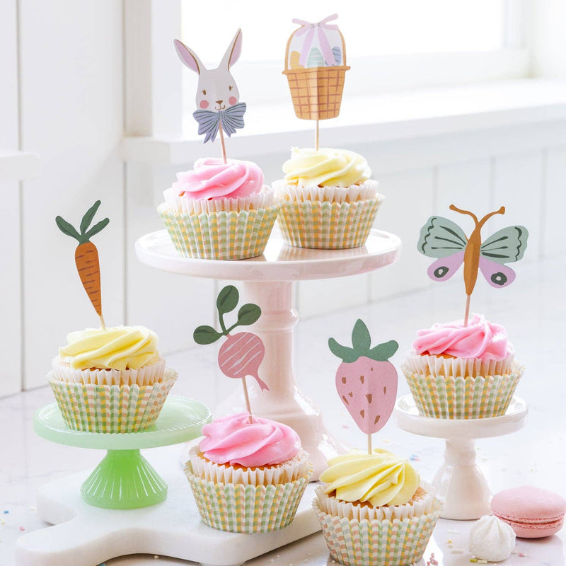 A tiered cake stand displaying cupcakes with pink and yellow frosting, decorated with bunny, carrot, and strawberry toppers from our Easter party supplies.