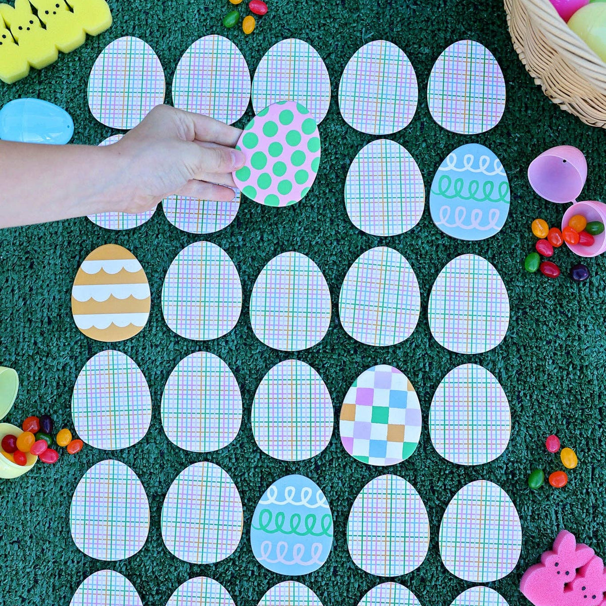 A lifestyle shot of a child's hand playing the Easter egg hunt memory match game on a green lawn surrounded by plastic eggs and jelly beans.