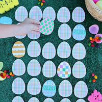 A lifestyle shot of a child's hand playing the Easter egg hunt memory match game on a green lawn surrounded by plastic eggs and jelly beans.
