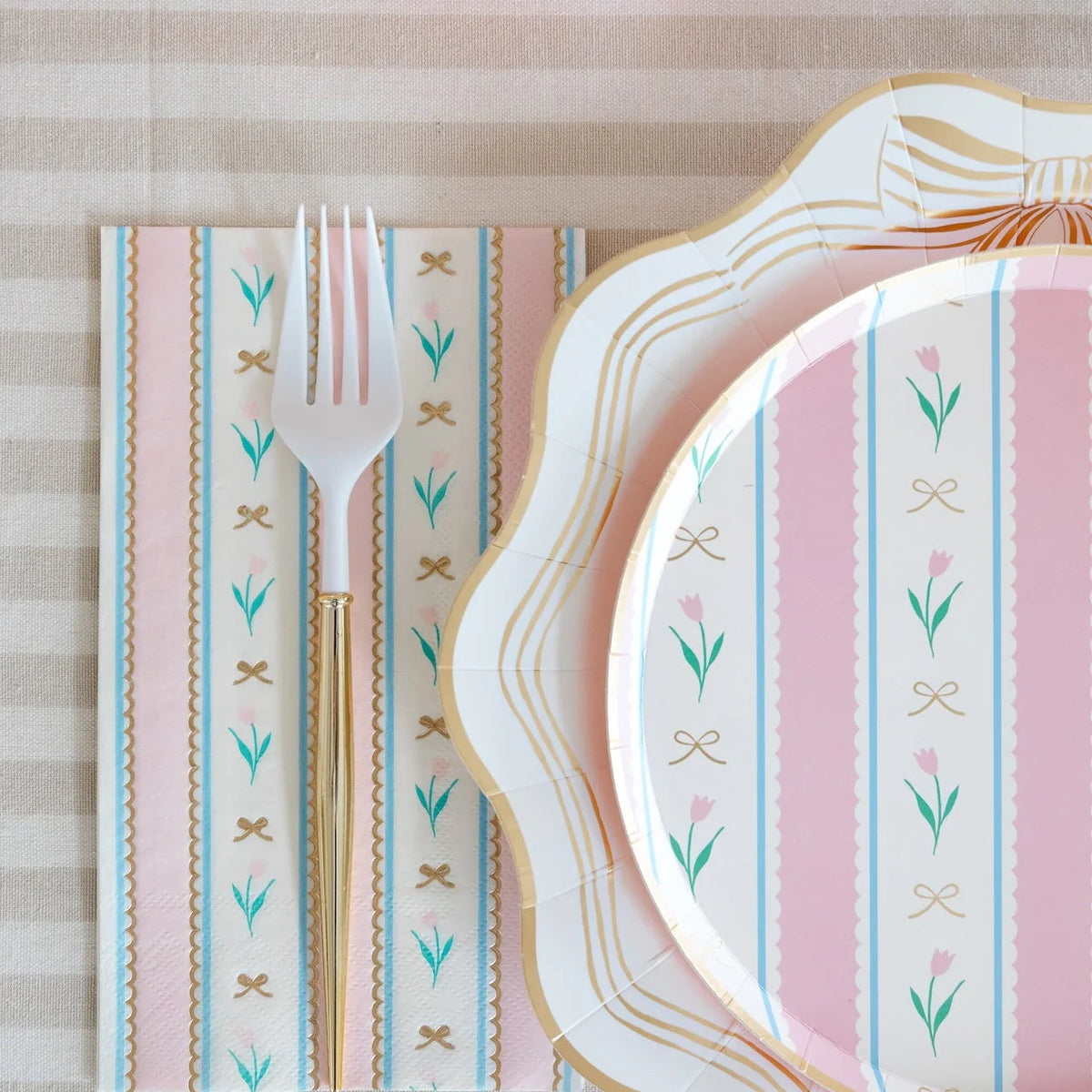 Pink and blue striped Easter plate stacked on scalloped dinner plate with matching napkin, white fork and gold cutlery on linen tablecloth