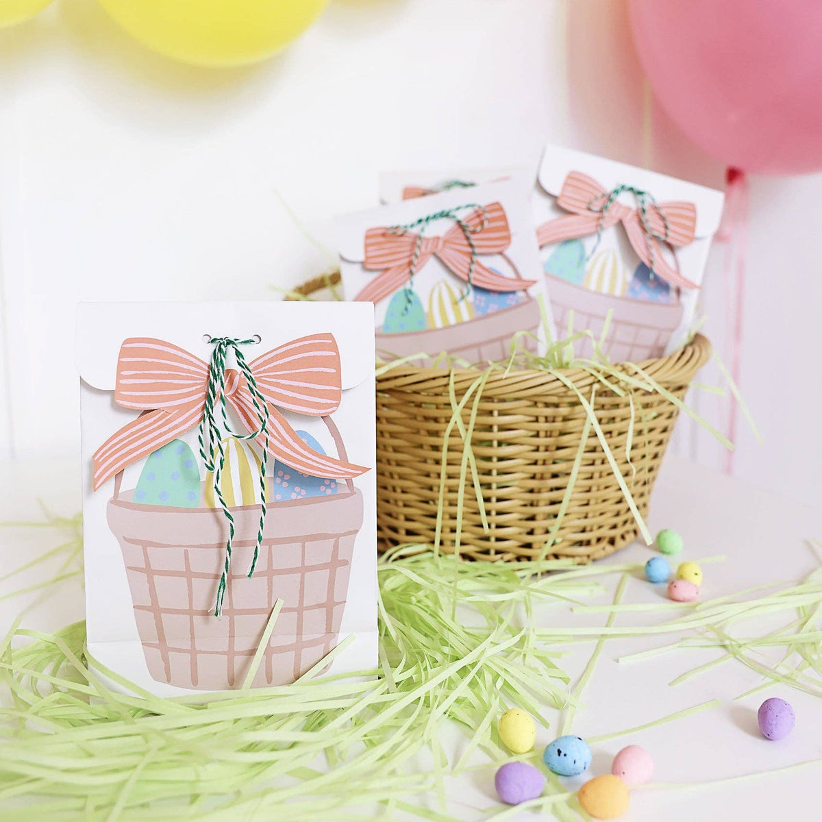 A lifestyle shot of three white paper treat bags featuring an illustrated Easter basket with a pink bow, sitting in a wicker basket with green paper grass and pastel candy.