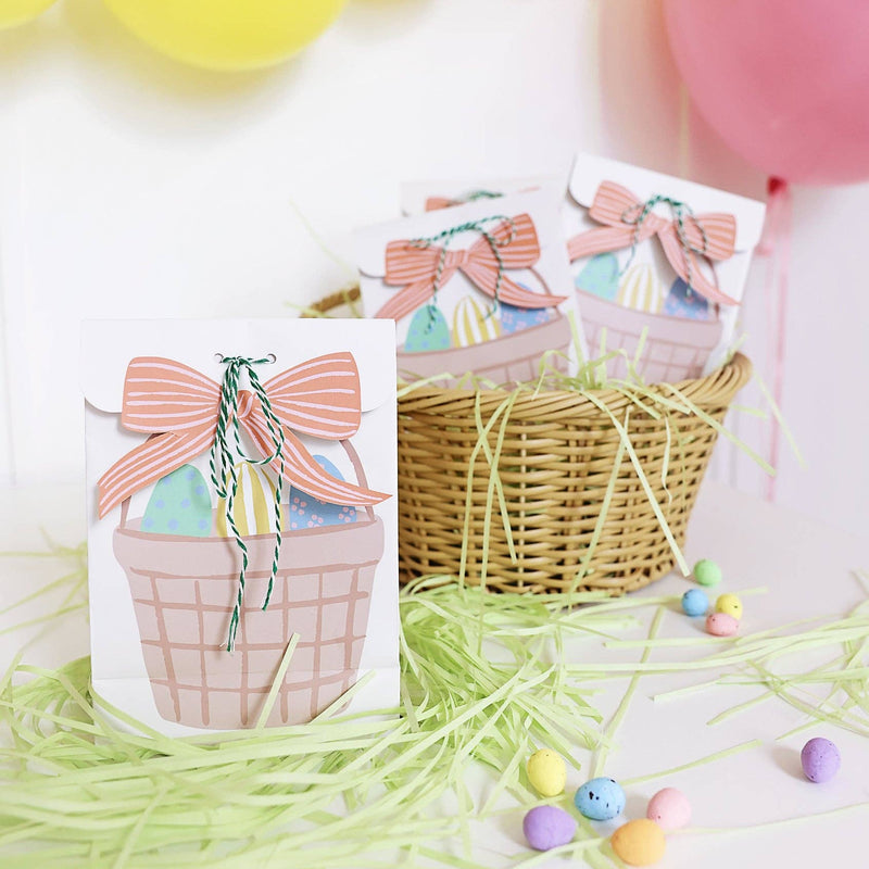A lifestyle shot of three white paper treat bags featuring an illustrated Easter basket with a pink bow, sitting in a wicker basket with green paper grass and pastel candy.