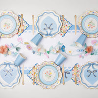 Overhead spring party tablescape featuring multicolor floral dinner plates, blue bow napkins, blue cups, and floral garland centerpiece