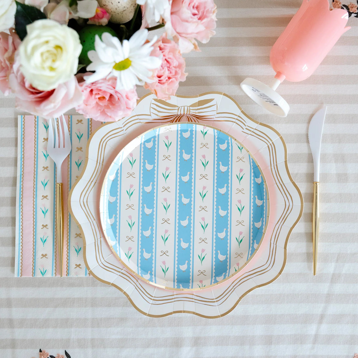 Overhead Easter place setting with blue goose striped plate on scalloped dinner plate, striped napkin, gold cutlery and pink roses