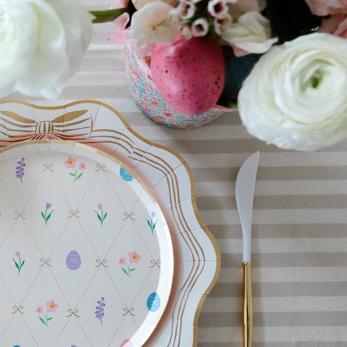 Close-up of pastel Easter small plate with gold bow and floral details layered on scalloped dinner plate with gold cutlery and white ranunculus