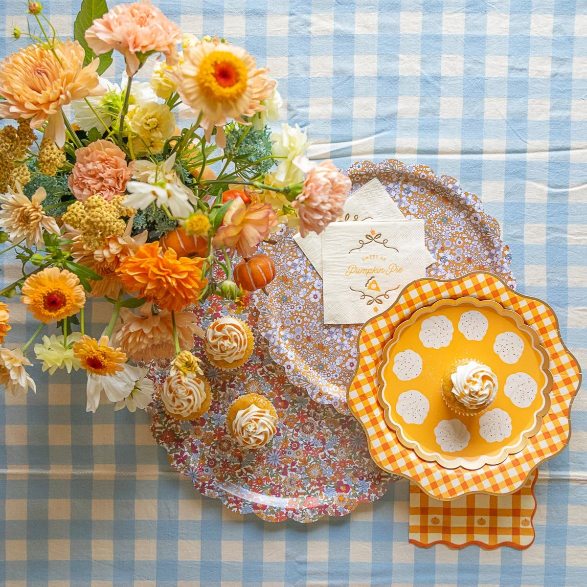 Top-down shot of blue gingham table with floral trays, cupcake, and Thanksgiving napkins in orange plaid.