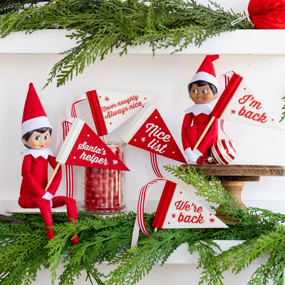 Two Elf on the Shelf dolls holding mini felt pennant flags with holiday sayings on a decorated shelf.