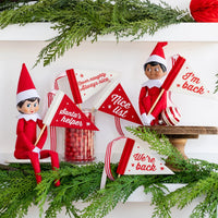 Two Elf on the Shelf dolls holding mini felt pennant flags with holiday sayings on a decorated shelf.