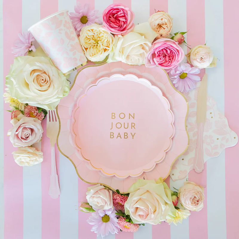 Overhead view of Bonjour Baby plates, napkins and cup arranged on pink‑striped runner with fresh roses and daisies.
