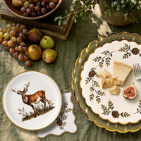 Overhead flat-lay with the stag plate, a pinecone plate with cheese and figs, grapes, pears, and greenery on an olive cloth.