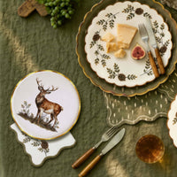 Overhead flat-lay with the evergreen cocktail napkin, a stag plate, a pinecone plate with cheese and figs, and greenery on an olive cloth.