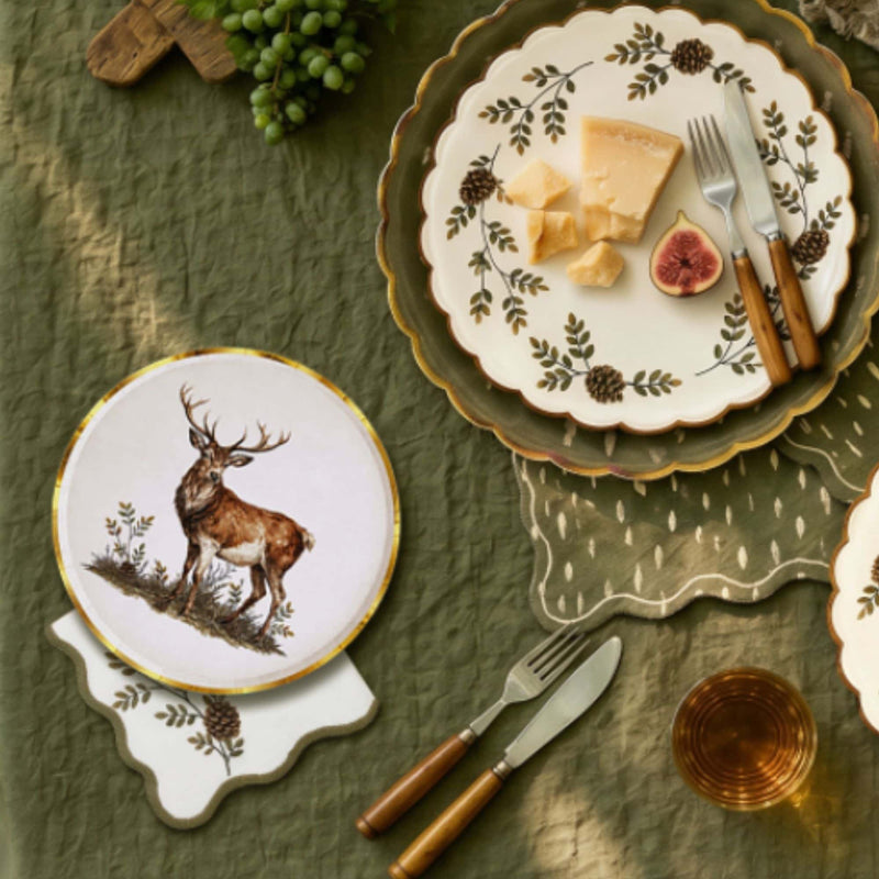 Overhead flat-lay with the evergreen cocktail napkin, a stag plate, a pinecone plate with cheese and figs, and greenery on an olive cloth.