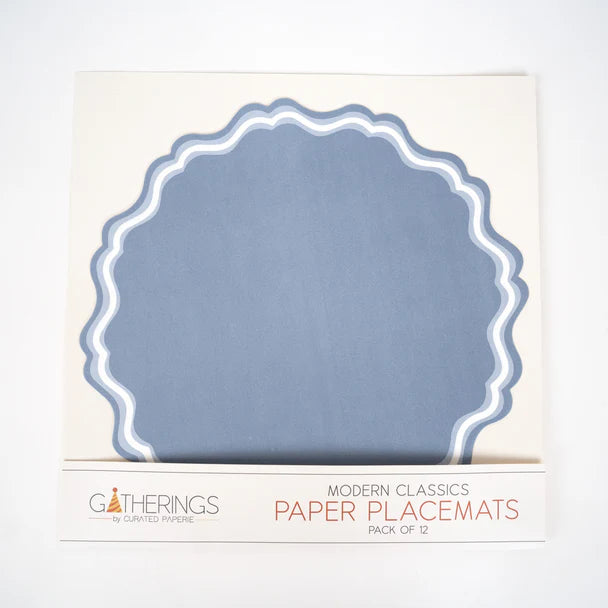 Front of retail box for paper placemats. Box has cream background with a rounded scalloped placemat visible through a window showing dusty blue paper with double-wave white and light blue border. Lower label reads "Gatherings by Curated Paperie / Modern Classics / Paper Placemats / Pack of 12."