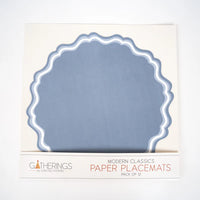 Front of retail box for paper placemats. Box has cream background with a rounded scalloped placemat visible through a window showing dusty blue paper with double-wave white and light blue border. Lower label reads "Gatherings by Curated Paperie / Modern Classics / Paper Placemats / Pack of 12."