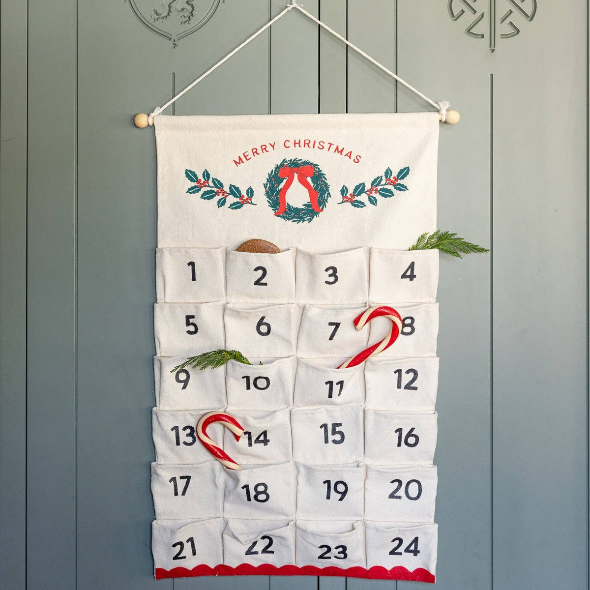 Fabric advent calendar hanging on green paneled door with candy canes and greenery in pockets