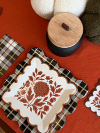 Fall tablescape with rust floral square paper plates, white and green knit pumpkins, and a wood-lidded candle on a rust orange tablecloth